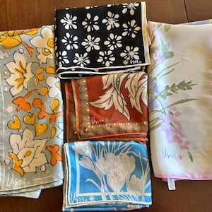 May include: A collection of silk scarves in various colors and floral designs. The scarves include a black one with white flowers, a rust-colored one with cream-colored leaves, and a light blue one with white flowers. Each scarf has a signature.