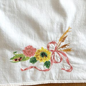 Puede incluir: Un tejido de tela blanca con un colorido motivo floral bordado. El diseño incluye un girasol amarillo, una flor rosa, hojas verdes y espigas de trigo doradas, todo ello atado con una cinta rosa fluida y una pequeña vaina verde.