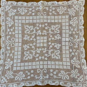 May include: A square white lace doily featuring an intricate openwork pattern of floral motifs and leaves. The design includes a central grid section surrounded by a decorative border with scalloped edges and repeating leaf-like shapes throughout.