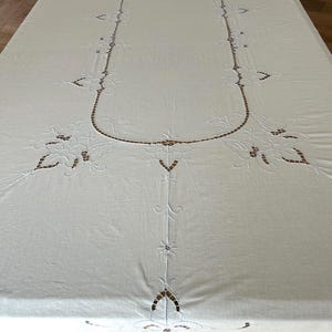 Puede incluir: Un mantel rectangular blanco con intrincados bordados calados. El diseño incluye un borde ovalado central con motivos florales en las esquinas y los lados, acentuado por un delicado trabajo de aguja y patrones calados en toda la tela.