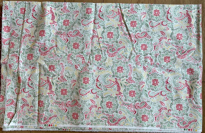 May include: A rectangular piece of fabric featuring a repeating pattern of red, green, and yellow paisley and floral motifs on a white background. The design includes intricate geometric shapes and swirling lines, with a finished edge at the bottom.