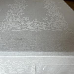 May include: A white rectangular tablecloth with an ornate, repeating floral pattern around the edges. The tablecloth is made of a textured fabric and is spread across a wooden table.