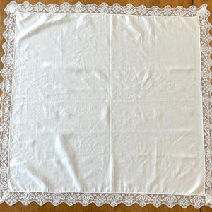 May include: A square white fabric napkin featuring a delicate, intricate lace border with a scalloped edge. The center of the napkin is made of smooth, plain white fabric, while the perimeter is framed by a detailed, repeating floral lace pattern.
