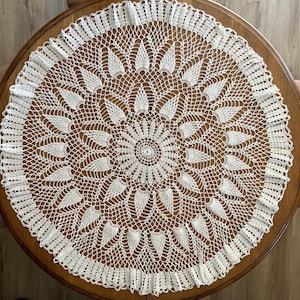 May include: A round white crochet doily featuring a central circular pattern that radiates outward into a series of leaf-shaped motifs, finished with a delicate scalloped edge and an intricate openwork mesh design throughout the entire piece.