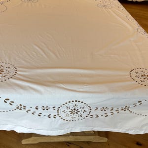 May include: White rectangular tablecloth with intricate cut-out floral and circular designs along the edges. The tablecloth is draped over a table, showcasing the detailed patterns and texture. The fabric appears to be cotton or linen.