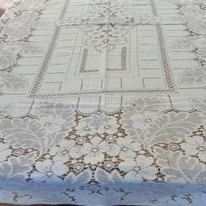 May include: A rectangular white lace tablecloth featuring an intricate floral pattern along the borders and a central geometric design with openwork detailing. The fabric displays a delicate, semi-sheer texture with a repeating leaf motif.
