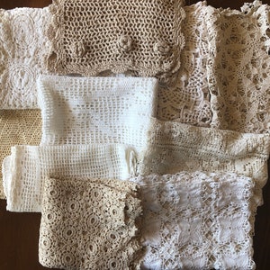 May include: A collection of handmade crochet doilies and lace pieces in various off-white and beige tones. The textiles feature intricate patterns, including floral and geometric designs. The items are arranged on a dark brown surface, showcasing the delicate textures.