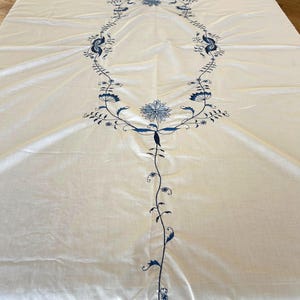 Puede incluir: Un mantel rectangular blanco con un motivo floral central bordado en azul. El diseño consiste en un marco ovalado alargado de enredaderas y hojas, acentuado con flores detalladas en la parte superior e inferior, que se extiende hacia abajo.