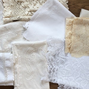 May include: Assortment of vintage white and cream-colored linens, including lace, embroidered handkerchiefs, and decorative edging. The textiles are arranged on a wooden surface, showcasing various textures and patterns.