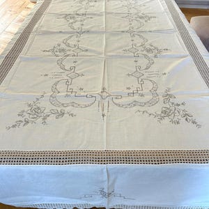 May include: A rectangular off-white tablecloth featuring intricate gray embroidered floral patterns and geometric lines. The edges are finished with a wide, decorative crocheted lace border that adds texture to the overall design.