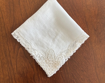 May include: A square white handkerchief featuring a delicate off-white lace border. The lace trim displays an intricate, repeating floral and geometric pattern along two edges, meeting at a pointed corner with a detailed, ornate lace motif.