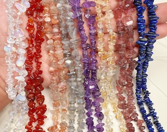 May include: Assortment of chip gemstone strands in various colors, including clear, red, orange, gray, purple, yellow, and blue. The strands are laid out in a row on a hand.