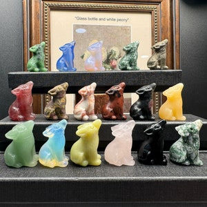 Puede incluir: Una colección de figuras de lobos de cristal talladas en varios colores, incluyendo verde, azul, rosa y amarillo. Los lobos están en una pose de aullido, exhibidos en un estante escalonado. Una imagen enmarcada está en el fondo.