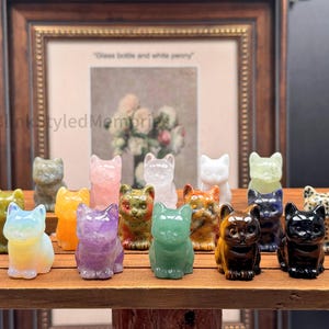 Puede incluir: Una colección de pequeñas figuras de gato de piedras preciosas en varios colores, incluyendo verde, morado, naranja y negro. Los gatos se exhiben en una estantería de madera, con una imagen enmarcada en el fondo.
