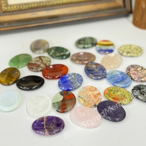 Peut inclure: Assortiment de pierres polies de forme ovale dans diff&eacute;rentes couleurs et motifs, notamment vert, rouge, bleu, marron, blanc et violet. Les pierres sont dispos&eacute;es sur une surface blanche.