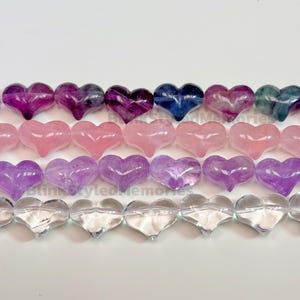 Puede incluir: Filas de cuentas en forma de corazón en varios colores, incluyendo morado, rosa y transparente. Las cuentas están dispuestas en líneas horizontales, mostrando sus superficies pulidas y variaciones de color únicas. Ideal para bisutería.