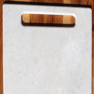 Op de afbeelding: Een rechthoekige, crèmekleurige snijplank met een ingebouwde handgreep. De plank heeft een gespikkeld patroon en ligt op een houten oppervlak. Dit keukenartikel is ontworpen voor het bereiden van voedsel.