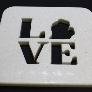 Op de afbeelding: Crèmekleurige vierkante onderzetter met het woord "LOVE" uitgesneden, met een staatsomtrek in plaats van de "O". De onderzetter heeft een gespikkelde textuur en afgeronde hoeken, tegen een zwarte achtergrond.