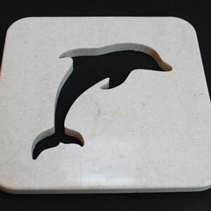 Peut inclure: Un dessous de verre carré beige clair avec un motif de dauphin découpé. Le dauphin saute, le corps arqué. Le dessous de verre a des coins arrondis et une surface lisse. Le fond est noir.