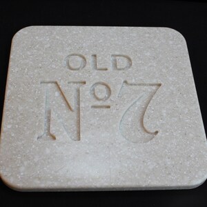 Może przedstawiać: Kwadratowa, nakrapiana, kremowa podstawka z zaokrąglonymi rogami. Na powierzchni wygrawerowano słowa "OLD" i "Nº 7". Podstawka wykonana jest z materiału przypominającego kamień.
