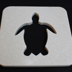 May include: A square, off-white coaster with a speckled texture. The center features a cutout in the shape of a sea turtle. The coaster is designed to protect surfaces from drink condensation and heat.