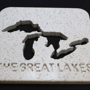 Puede incluir: Un posavasos cuadrado blanco moteado con un mapa tallado de los Grandes Lagos. Las palabras "THE GREAT LAKES" están talladas en la parte inferior del posavasos. Los lagos son de color gris oscuro.
