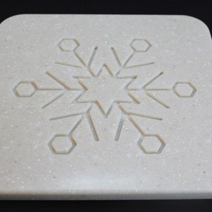 Pode incluir: Uma base para copos quadrada, de cor branco-sujo, com um desenho de floco de neve gravado na superfície. O floco de neve apresenta uma forma de estrela central com linhas radiais e formas hexagonais. A base tem cantos arredondados.