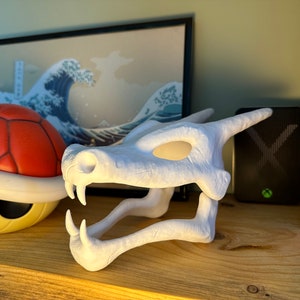 May include: White plastic dragon skull figurine with a detailed design. The skull is open-mouthed with sharp teeth and has a hollow eye socket.