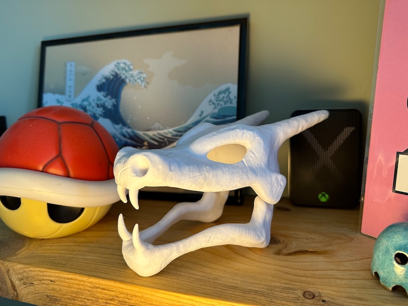 May include: White plastic dragon skull figurine with a detailed design. The skull is open-mouthed with sharp teeth and has a hollow eye socket.