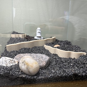 May include: A small, white figurine of a person's head with a black face, set in a black gravel terrarium with a beige, wavy border. There are two white rocks and a small, brown shell in the gravel.