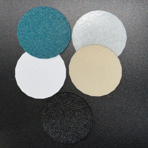 May include: Five circular, textured pads in varying colors: teal, white, beige, black, and a light white. The pads are arranged in a cluster, with the black pad at the bottom. The surfaces have a slightly glittery appearance.