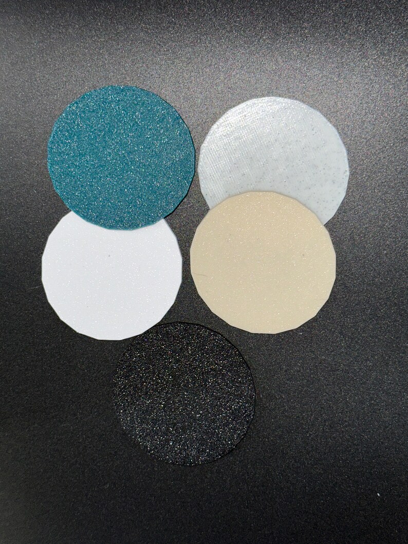 May include: Five circular, textured pads in varying colors: teal, white, beige, black, and a light white. The pads are arranged in a cluster, with the black pad at the bottom. The surfaces have a slightly glittery appearance.