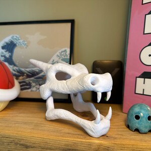 May include: White plastic 3D printed model of a dragon skull. The skull is open-mouthed and has a textured surface.