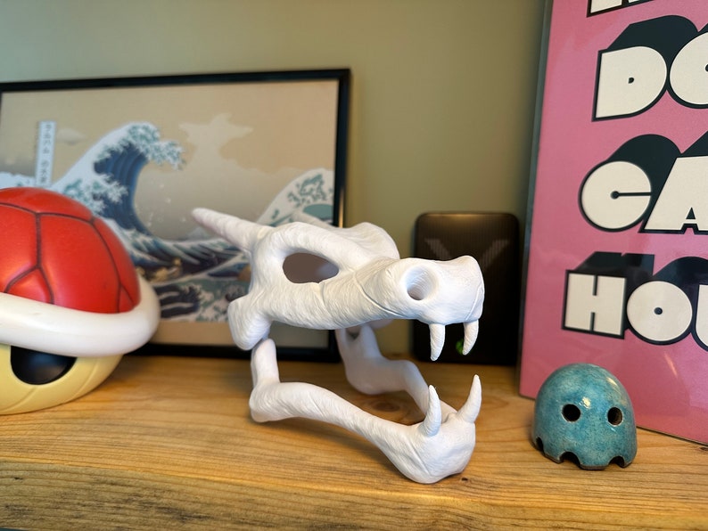 May include: White plastic 3D printed model of a dragon skull. The skull is open-mouthed and has a textured surface.