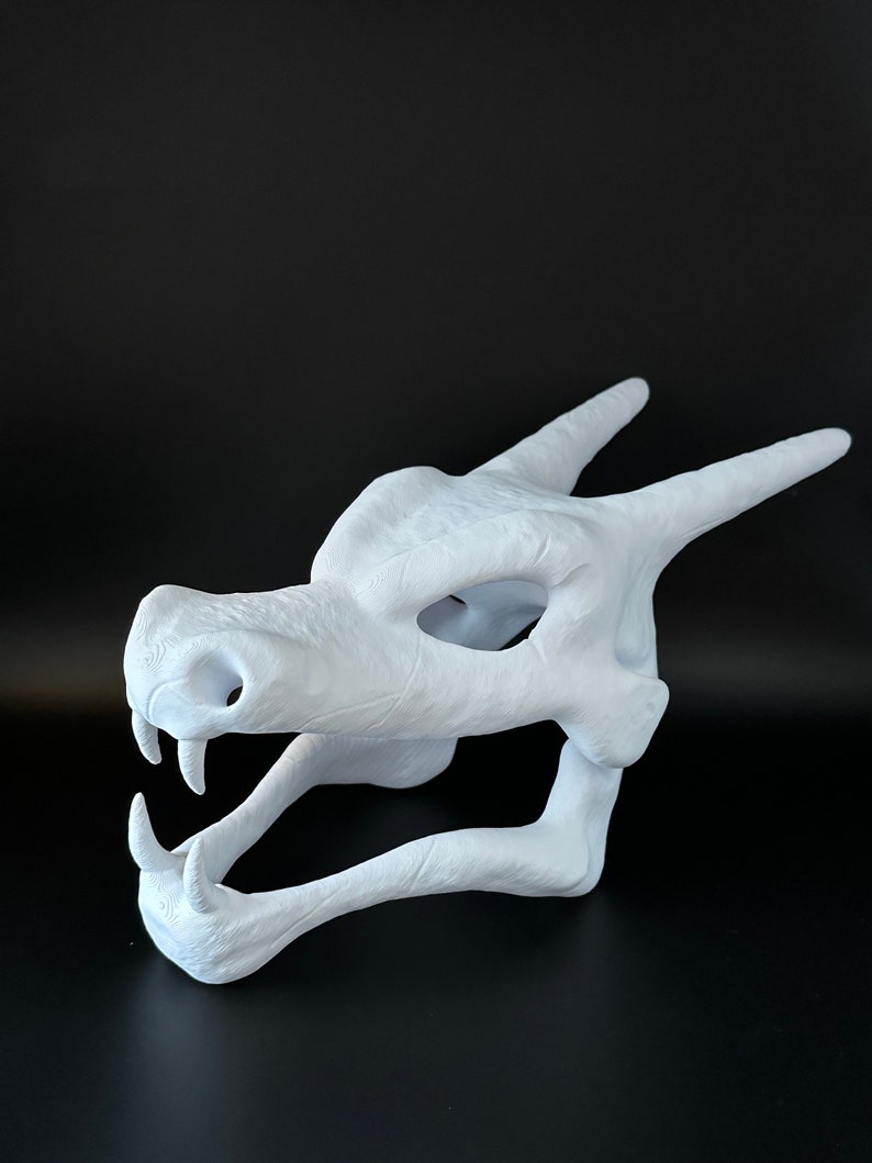 May include: A white 3D printed dragon skull mask. The mask is open-mouthed and has a detailed design.
