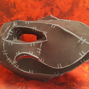 May include: A black paper mask with white stitching details. The mask has a large eye hole and a curved shape.