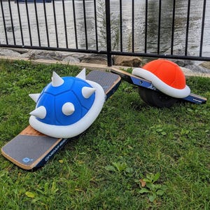 May include: Two Onewheel electric skateboards with custom-made Mario Kart-themed shells. One features a blue shell with white spikes, the other an orange shell. The boards are on green grass with a city and bridge in the background.