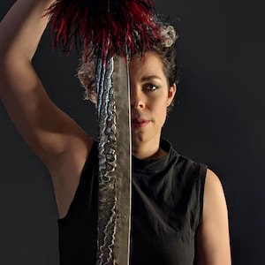 May include: A decorative sword with a textured, silver blade and a handle adorned with red and black feathers. The sword is held up in front of a person wearing a black sleeveless top.