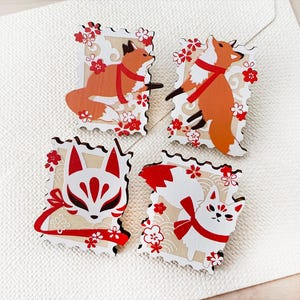 May include: Four rectangular wooden pins with scalloped edges, each featuring a fox-themed illustration. The designs include two orange foxes with red scarves, a white fox mask with red markings, and a white fox with a red scarf and red tail tip.