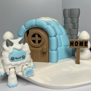 May include: A whimsical winter scene featuring a blue igloo with a wooden door, a sign that says "HOME", and a friendly yeti figure. The scene is set on a white base, with a gray chimney and snowballs.