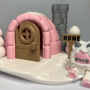 Könnte beinhalten: Ein skurriles Diorama mit einem rosa Iglu mit Holztür, einem Schneemann und einem Schild mit der Aufschrift "HOME". Eine weiße, pelzige Kreatur mit rosa Akzenten ist ebenfalls zu sehen. Die Szene ist auf einer weißen Basis aufgebaut.