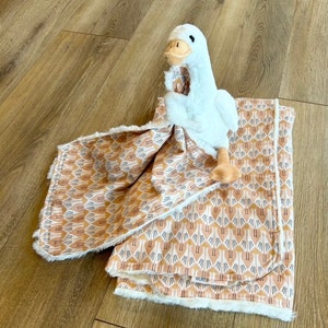 May include: A white plush goose toy is lying on a brown and white patterned baby blanket. The blanket has a white fleece backing.