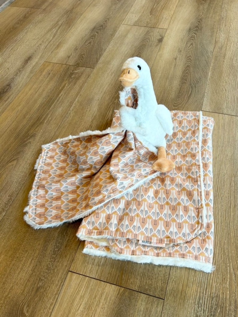 May include: A white plush goose toy is lying on a brown and white patterned baby blanket. The blanket has a white fleece backing.