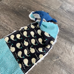 May include: A blue whale plush toy is nestled in a soft, white-trimmed baby blanket. The blanket features a patchwork design with a black fabric printed with white whales and a light blue fabric with white polka dots.