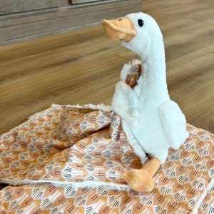 May include: A white plush goose toy is lying on a pink and white patterned blanket. The blanket has a repeating pattern of stylized geese.