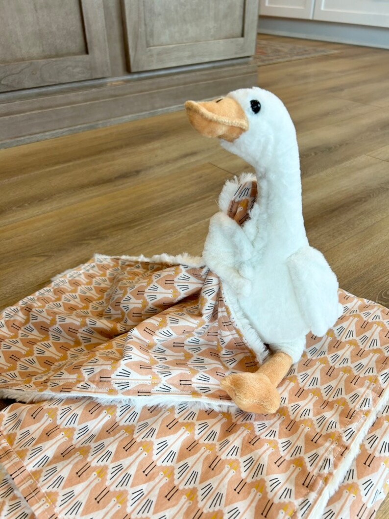 May include: A white plush goose toy is lying on a pink and white patterned blanket. The blanket has a repeating pattern of stylized geese.