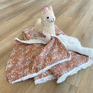 May include: A brown and white floral patterned baby blanket with a white faux fur border. A white plush llama toy is sitting on top of the blanket.
