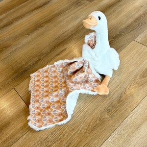 May include: A white plush goose toy with a brown beak and eyes, sitting on a brown and white patterned baby blanket.