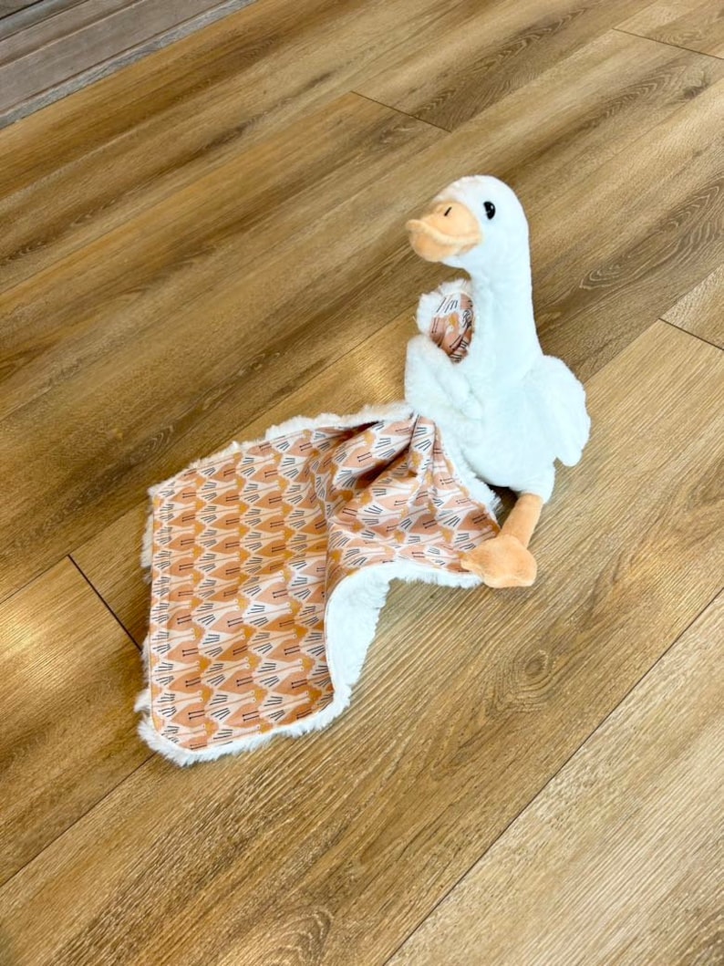 May include: A white plush goose toy with a brown beak and eyes, sitting on a brown and white patterned baby blanket.