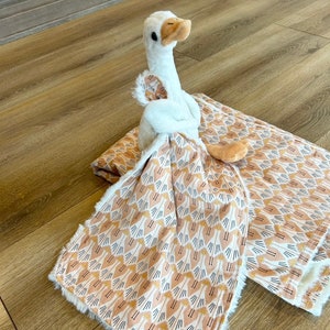 May include: A white plush goose toy is lying on a brown and white patterned baby blanket. The blanket has a soft, fuzzy texture and features a repeating pattern of small geese.